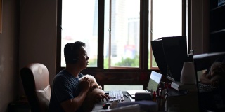 一位亚洲华人中年男子在网上与朋友、家人和商业伙伴聊天、聊天，一边在家里办公室的电脑屏幕上看着摄像头聊天，一边带着他的玩具狮子狗
