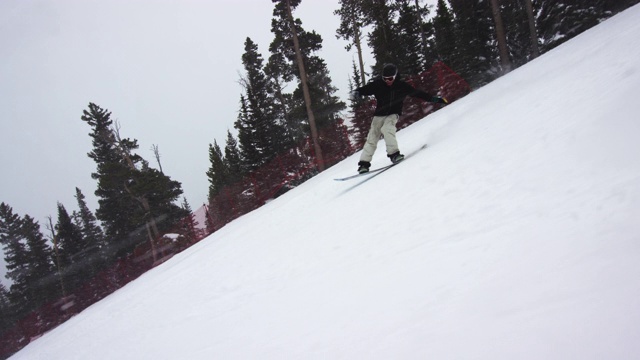 慢动作拍摄的滑雪板穿着完整的冬季装备停止在滑雪跑后做了一个“BS黄油”的伎俩被森林包围在博尔德附近的埃尔朵拉滑雪度假村，科罗拉多州在一个下雪的，阴天，雪遍布周围