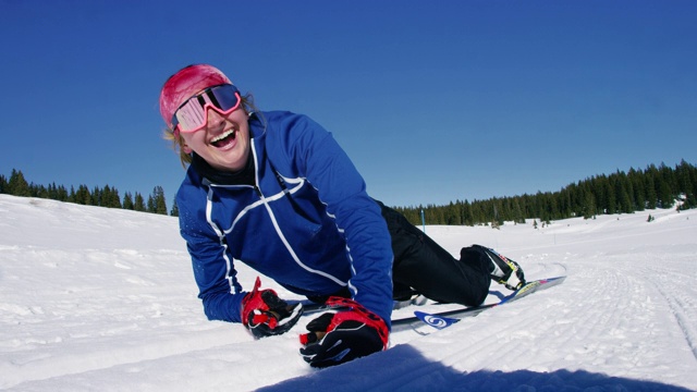慢动作镜头的一个年轻女子在她的二十多岁的越野滑雪，摔倒在雪地上，并笑着看着相机