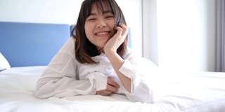 美丽的亚洲女人微笑幸福的床上，她躺在卧室的毯子下，舒适舒适的休息在酒店房间，人们健康的生活方式在早上