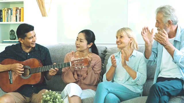 一群老年人在客厅里玩吉他开派对。白人，亚洲人，泰国人坐在家里的沙发上。退休生活方式的概念。前面的镜头。