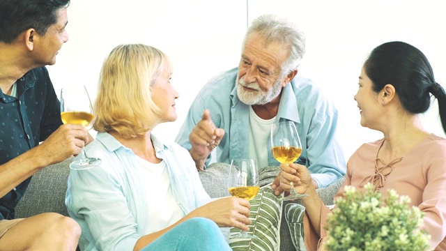 一个老人在客厅里喝着白葡萄酒和妻子聊天。白人，亚洲人，泰国人坐在家里的沙发上。退休生活方式的概念。