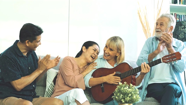 一位年长的女士和她的朋友在客厅玩吉他。白人，亚洲人，泰国人坐在家里的沙发上。退休生活方式的概念。