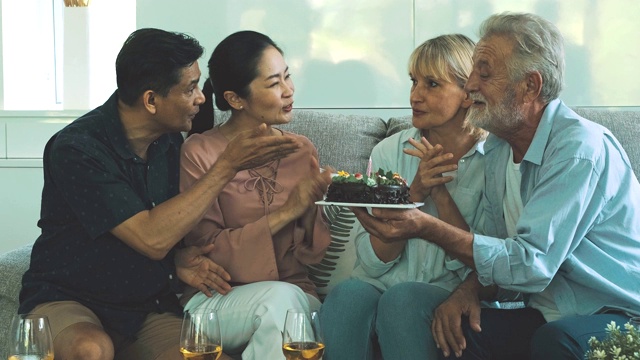 一群老年人在客厅举行生日聚会鼓掌。白人，亚洲人，泰国人坐在家里的沙发上。退休生活方式的概念。