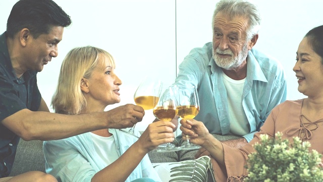 一群老年人在客厅聚会，喝着白葡萄酒，一起庆祝。白人，亚洲人，泰国人坐在家里的沙发上。退休生活方式的概念。