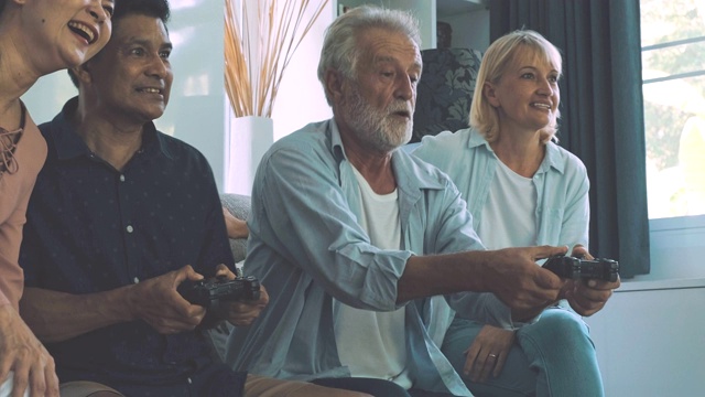 一群老年人在客厅玩电脑游戏，大获全胜。白人，亚洲人，泰国人坐在家里的沙发上。退休生活方式的概念。