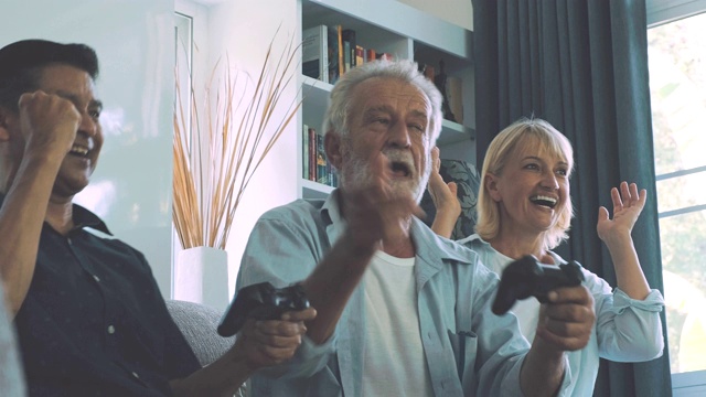 一群老年人在客厅玩电脑游戏，赢了又输。白人，亚洲人，泰国人坐在家里的沙发上。退休生活方式的概念。