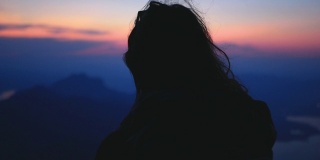 长棕色头发的徒步旅行者女孩在视点看日落在蓝色小时。风吹女人的头发在高山风景和湖泊。60帧/秒的慢镜头山峰