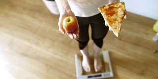 SLO MO拍摄了一个面目人非的女人一手拿着一片披萨，一手拿着一个苹果踩在体重秤上的照片。
