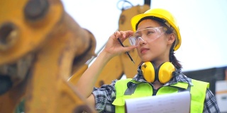 亚洲女工程师站在挖掘机的长柄勺旁，检查一个方案