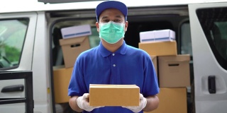 冠状病毒概念，蓝色快递英俊的亚洲男子拿着包裹纸盒与防护口罩和医用橡胶手套站在货车前面。4k分辨率和慢动作拍摄。