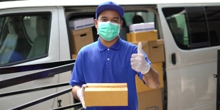 冠状病毒概念，蓝色快递英俊的亚洲男子拿着包裹纸盒，竖起大拇指，戴着防护面具和医用橡胶手套站在货车前面。慢动作镜头。