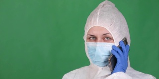 年轻的女医务人员在一个绿色的背景下讲电话，在一个白色的雾大衣防护面具和橡胶手套