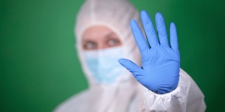 年轻的女医生穿着白色的鼠疫防护服，戴着蓝色的橡胶手套，在绿色的背景下显示停止的手势