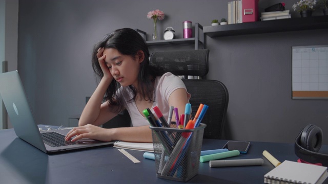 迷人的亚洲少女感到悲伤和沮丧后阅读和学习网上课程在她的笔记本电脑。在家上学的女孩坐在家里的客厅里，担心她的在线学习和教育考试成绩