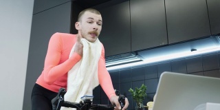英俊的男人做健身有氧运动智能自行车教练和擦脸与毛巾。男性运动员在自我隔离期间在家准备在线自行车比赛