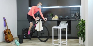 穿着自行车服的男子在室内智能自行车训练器上做有氧运动，在新冠肺炎隔离期间呆在家里。一个人骑着健身车，用毛巾擦脸