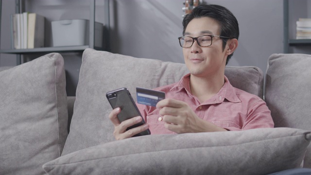 亚洲男人喜欢使用手机或智能手机进行网上购物，并持有信用卡支付他的订单。成年人坐在家里客厅的沙发上进行在线支付。购买，电子商务，网上商店和技术。