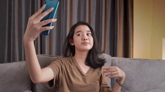 迷人的亚洲少女坐在客厅的沙发上用手机自拍。女孩喜欢在家里拍照。