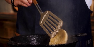 男性厨师油炸卡门贝尔奶酪在一个黑色的铁平底锅从两边与金属铲。资料片。在餐厅里准备食物的过程