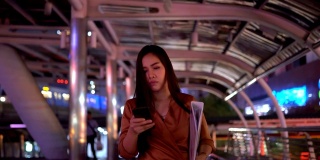 美丽年轻的亚洲女人走在夜晚的城市街道上使用智能手机