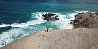 一架无人机拍摄到一对夫妇站在悬崖边欣赏风景。俯瞰印尼龙目岛丹戎安海滩。绿松石色的水。幸福和和睦