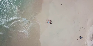 一对夫妇躺在印度尼西亚龙目岛粉红色海滩的沙滩上玩无人机。波浪轻轻地拍打着海岸。他们晒黑。快乐和粗心的时刻。
