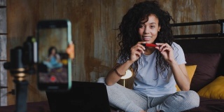 年轻的非洲裔美国女性化妆博主坐在家里的床上，用智能手机评论化妆产品，摄影