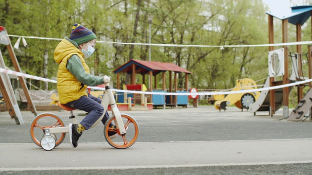 在因新冠肺炎疫情而关闭的游乐场附近，戴着防护面具的小男孩骑着跑步自行车