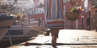 年轻女子提着花篮走在意大利庞扎岛港口的海滨。时尚色彩艳丽的礼服、裙子和帽子。