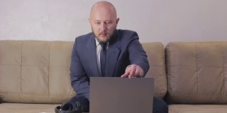 白种秃顶的年轻人，留着胡子，穿着西装，坐在沙发上，在家庭办公室的笔记本电脑上工作，在隔离时间