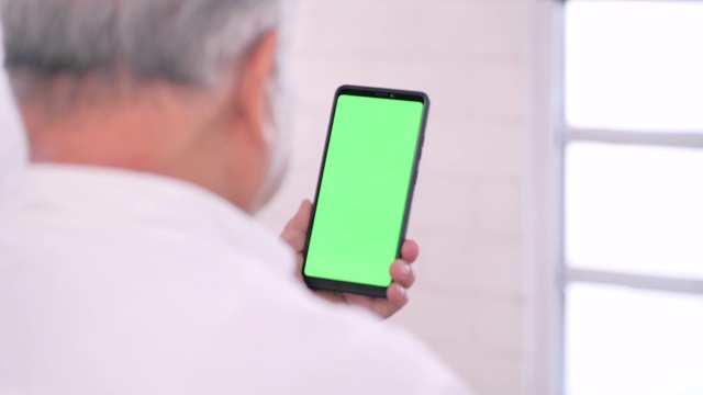 亚洲老爷爷在家看智能手机绿屏，用智能手机进行视频会议。家庭，高级卫生技术，医疗，退休，电子学习，医学和保健。绿色屏幕手持智能手机