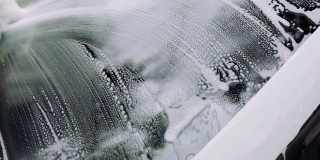 男人的手用海绵和泡沫擦洗汽车的挡风玻璃。汽车服务理念，