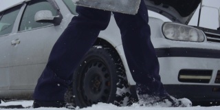冬天，人们从覆盖着积雪的地面上举起汽车电池，把它送到汽车上。一个在下雪时修理坏了的汽车。车辆故障或问题。引擎盖或引擎盖抬起