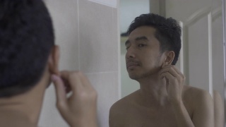年轻的亚洲男子在脸上涂霜看着镜子视频素材模板下载