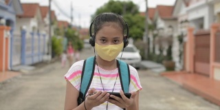 亚洲中学女生戴着防护面罩，戴着耳机，在住宅区行走。女青少年背包使用手机，而她是在回家的路上。