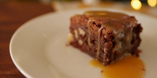 一块方形的巧克力布朗尼蛋糕放在一个盘子里，在一个木制的背景上，一张有黄色花朵的餐巾和一个旧的梅尔基奥勺子。金黄的咸味焦糖慢慢沥干。