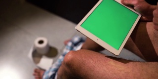 一名男子坐在马桶或壶上，手里拿着绿色屏幕的平板电脑的视频。向下滑动几次。地板上有一卷厕纸。