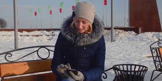 漂亮的女孩穿着暖和的衣服，戴着帽子，在冬天，坐在公园外面的长凳上打电话