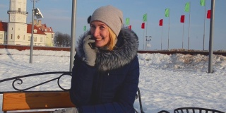 漂亮的女孩穿着暖和的衣服，戴着帽子，在冬天，坐在公园外面的长凳上打电话