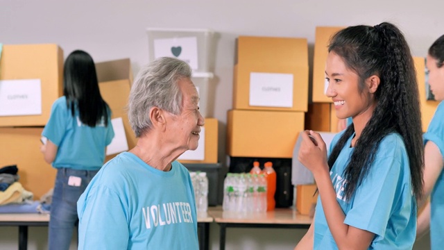 描绘70岁的亚洲祖母和年轻的黑人妇女看着相机，而一群朋友在为新冠病毒危机中的人们打包捐赠衣服。志愿服务
