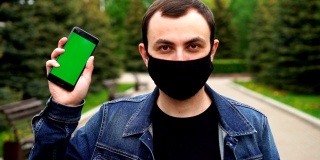 身着黑色医用口罩的男子在户外展示绿色屏幕的手机，背景为绿色