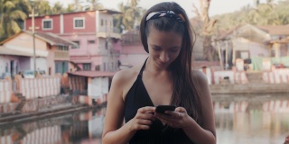 在城市背景下输入智能手机的年轻女性的户外肖像。长发女孩拿着手机在印度小镇看短信慢镜头。暑期技术交流