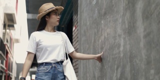 美丽的亚洲女人戴着一顶复古的帽子，拿着一个布袋，用手轻轻触摸着墙的背景，看着一条小街道享受着假期的夏天。