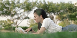 迷人的亚洲女人放松地读一本书，而躺在地板上的草地在户外的一个公园在美丽的日落。