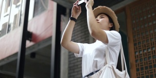 美丽的亚洲女人戴着软呢帽，站在老城区的一条小街上用智能手机拍照。快乐少年假日旅行生活方式。