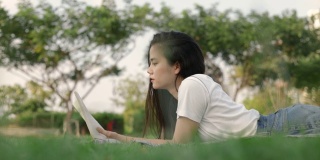 美丽的亚洲女人放松阅读一本书，而躺在地板上的草地户外在一个公园在美丽的日落。