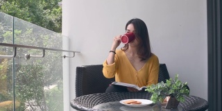 一位亚洲华裔美女中午在她的阳台上看书，放松，享受她的数字排毒时间，喝着咖啡，吃着饼干当点心