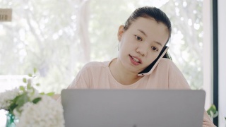 亚洲商务女性在家工作，桌上放着智能手机和笔记本电脑，心情愉快。社会距离的概念，以阻止冠状病毒疾病的传播。4 k UHD。缓慢的运动。视频素材模板下载