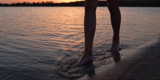男性的腿和脚沿着湖或池塘在黄昏的沙滩上行走。人们只能在岸上行动。背景是平静的水。120帧/秒的慢动作。夕阳下，一个人在海湾或小溪沐浴后散步